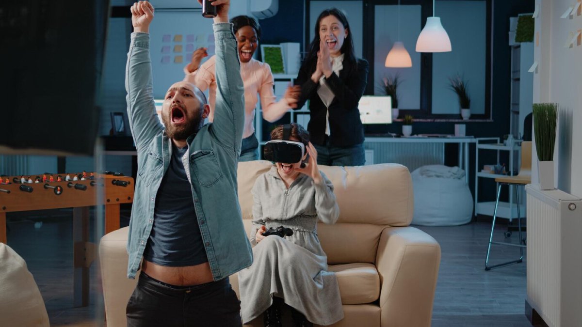 A group of friends celebrate joyfully in a cozy room, with one person cheering and others clapping in excitement. Gaming console on sofa.