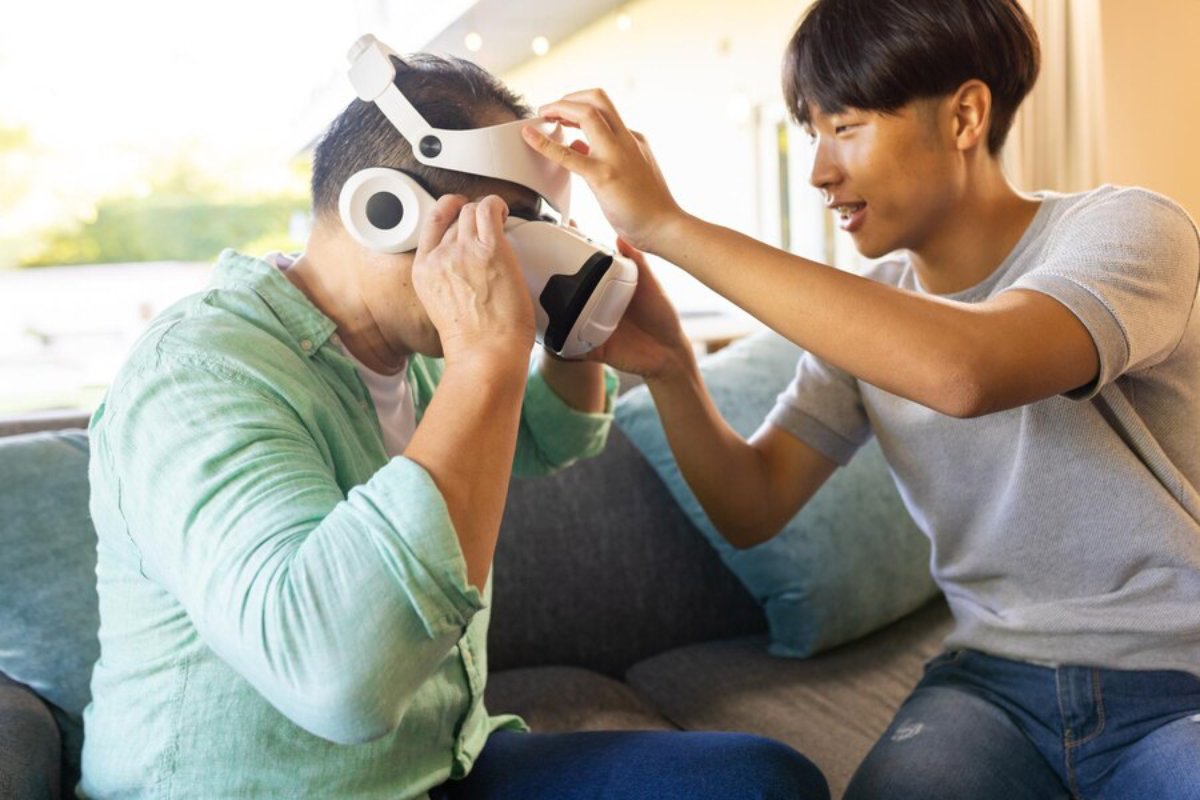 Two men on a couch, one adjusting a virtual reality headset on the other's head, smiling in a sunny living room.