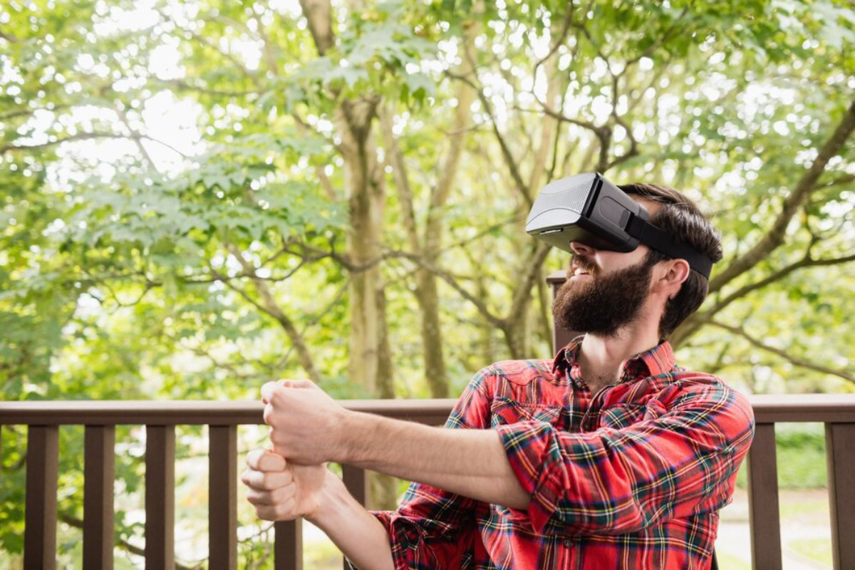 Bearded man wearing a VR headset, enjoying a simulation outdoors on a deck surrounded by green trees.