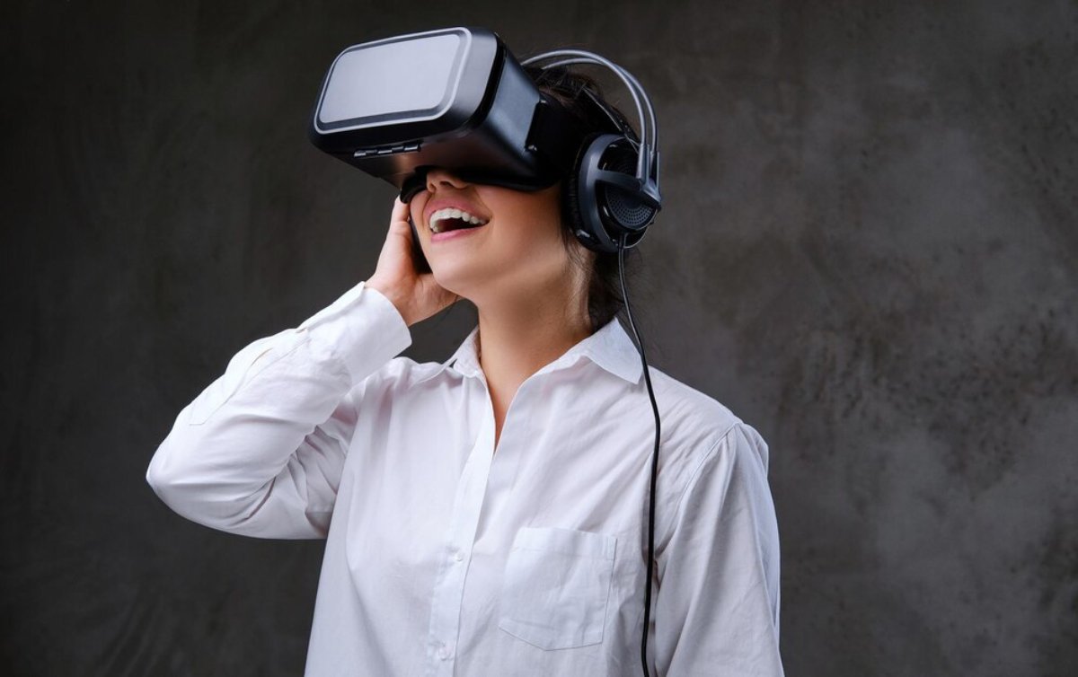 A woman in a white shirt wearing a VR headset and headphones, smiling and touching the headset.