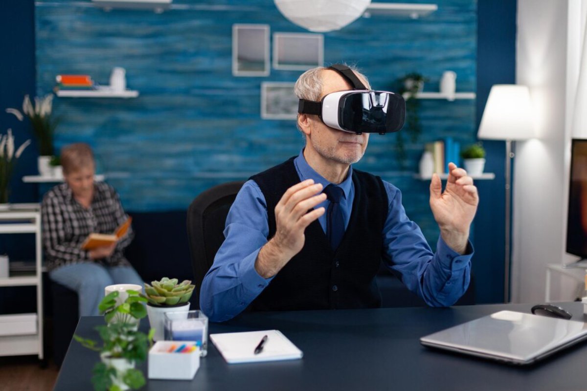 Elderly man using VR headset in a modern office with a woman reading in the background.