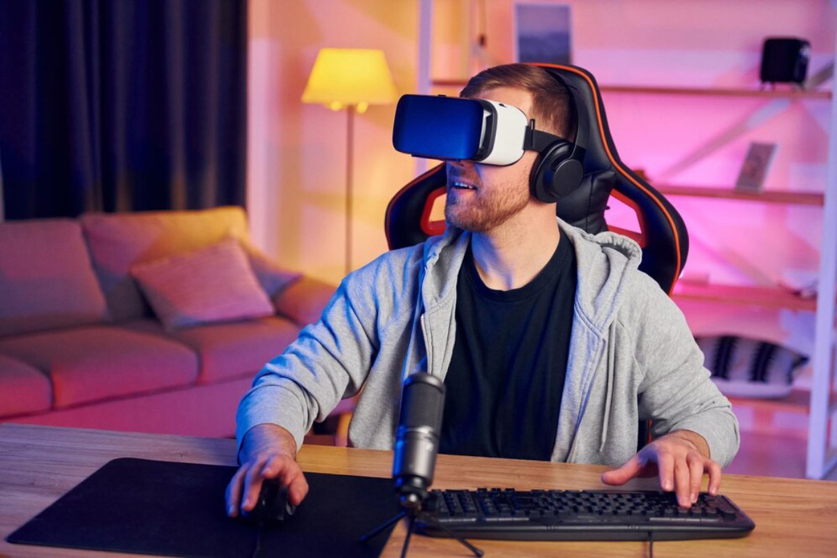 Man wearing a VR headset and headphones sits at a computer desk in a warmly lit room.