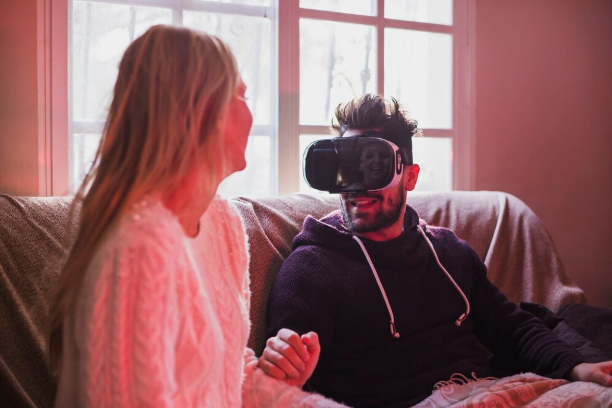 Man wearing VR headset sitting on couch beside a woman, both illuminated by warm light from a window.