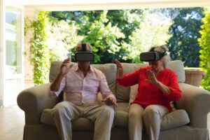 Elderly couple wearing virtual reality headsets on a sofa, gesturing with hands, surrounded by a bright room with garden view.