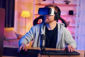Man wearing virtual reality headset sitting at a desk with microphone and computer in a colorful room.