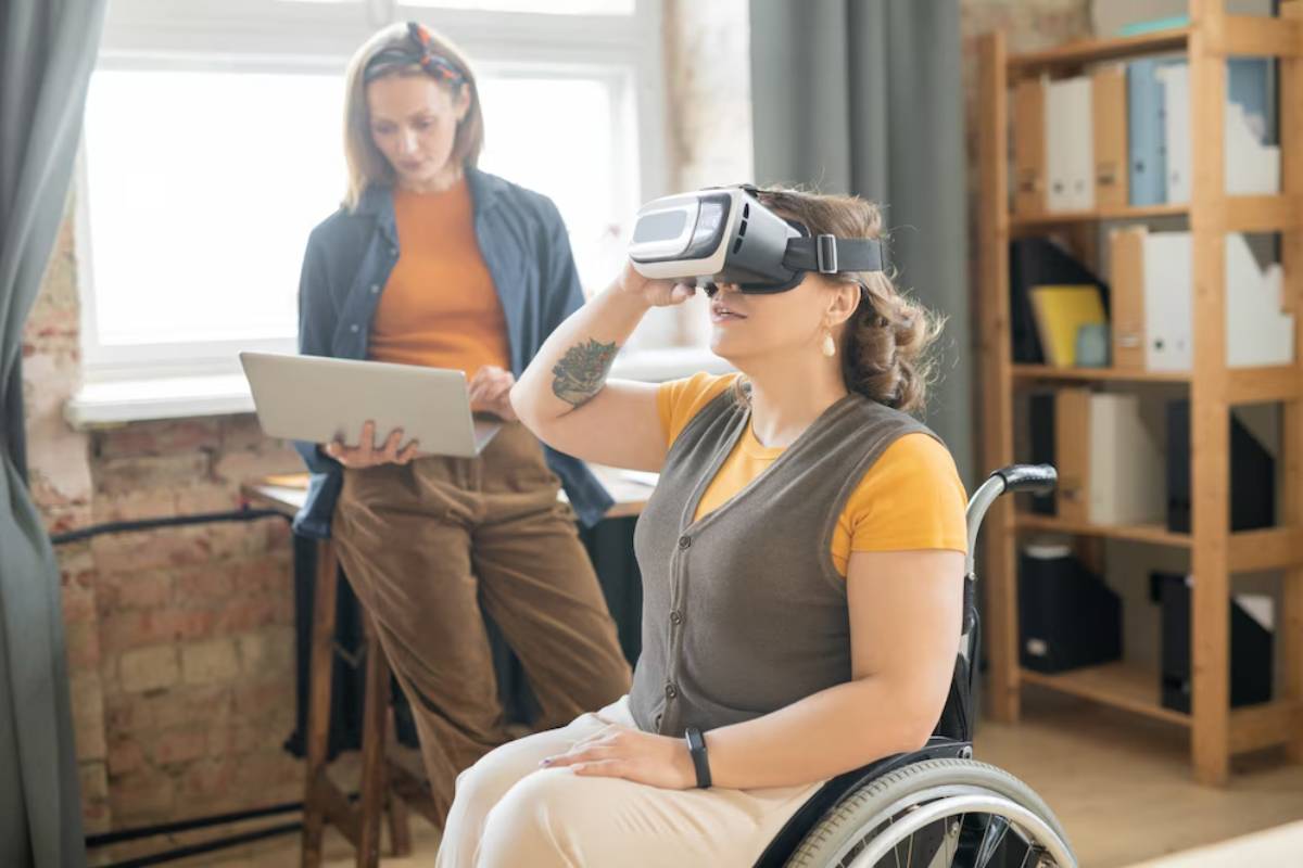 Woman in a wheelchair experiencing virtual reality, while another woman with a laptop observes in an office setting.