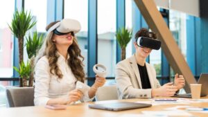 A woman and a man wearing virtual reality headsets sit at a table with plants, laptops, and design materials, engaged in a tech session.