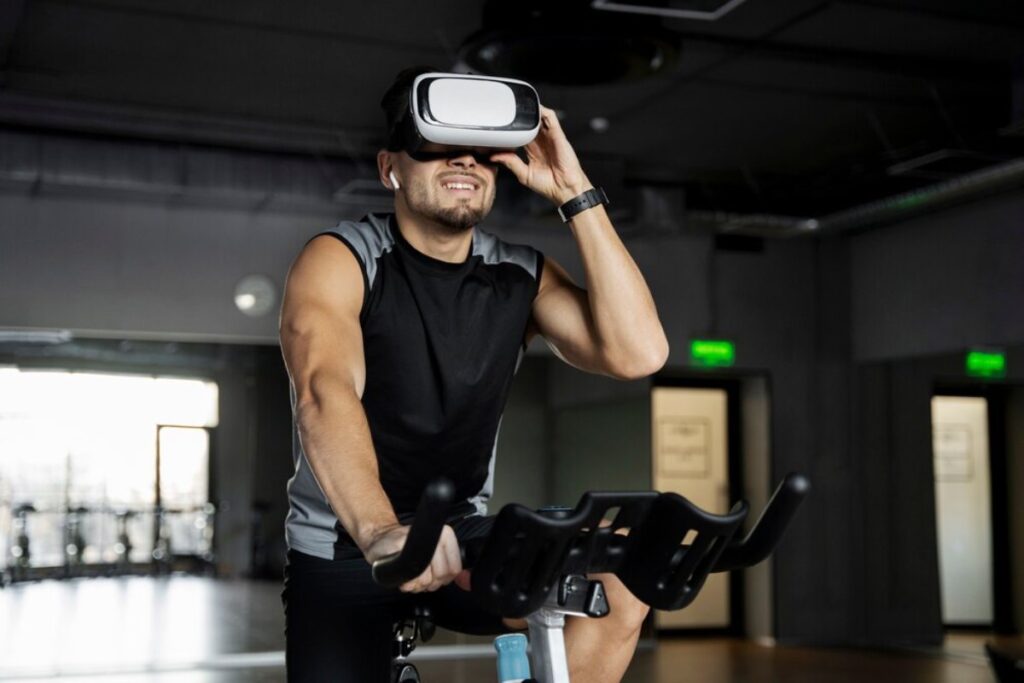 Man in black sleeveless shirt using virtual reality headset while riding a stationary bike in a gym.
