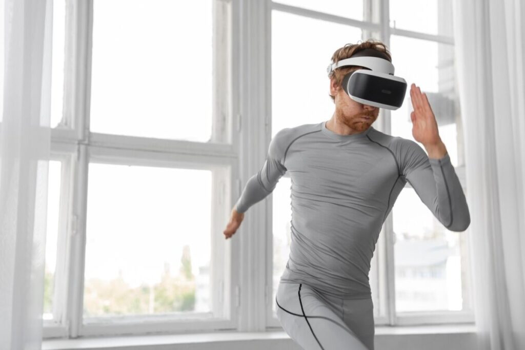 Man in gray athletic wear using a VR headset in a bright room with large windows.