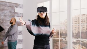 Woman using a virtual reality headset gestures with her hands, another person observes in a brightly lit room with large windows.