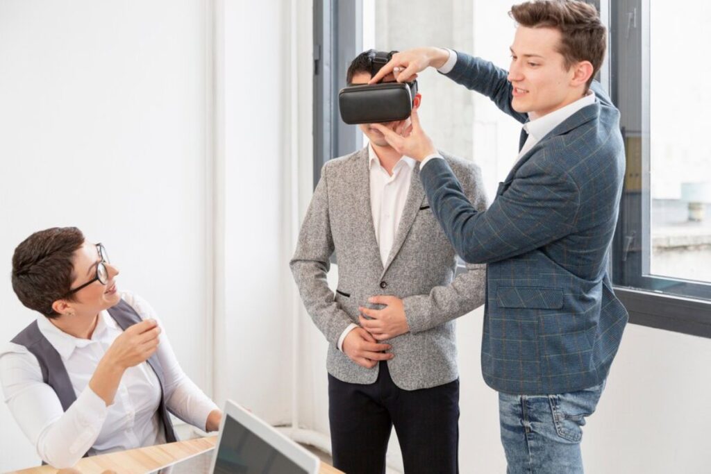 Two men and a woman in an office, one man helping another try on a virtual reality headset.