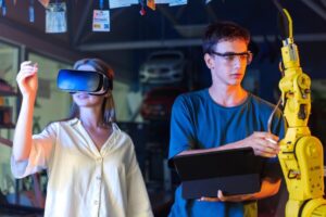A woman wearing VR goggles and a man with a laptop next to a robotic arm in a workshop.